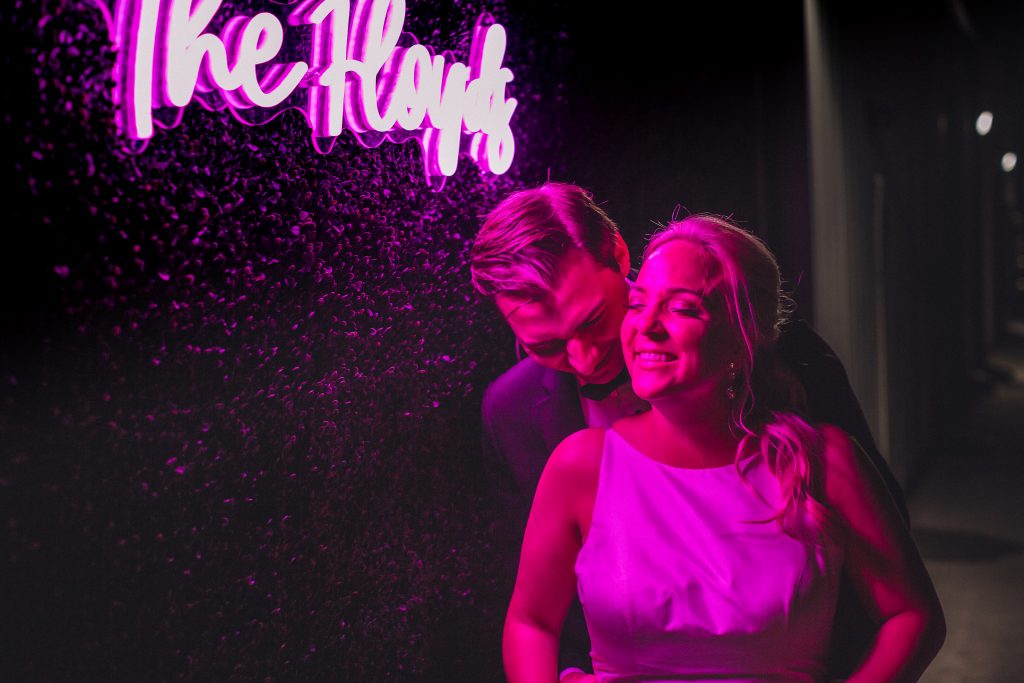 Bride and groom embrace under a pink neon sign that shows off their new shared last name.